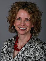 Smiling woman with curly hair wearing a patterned jacket and red necklace.