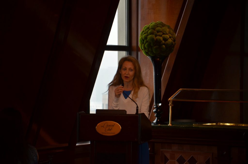 Woman speaking at a podium in a dimly lit room with large windows.