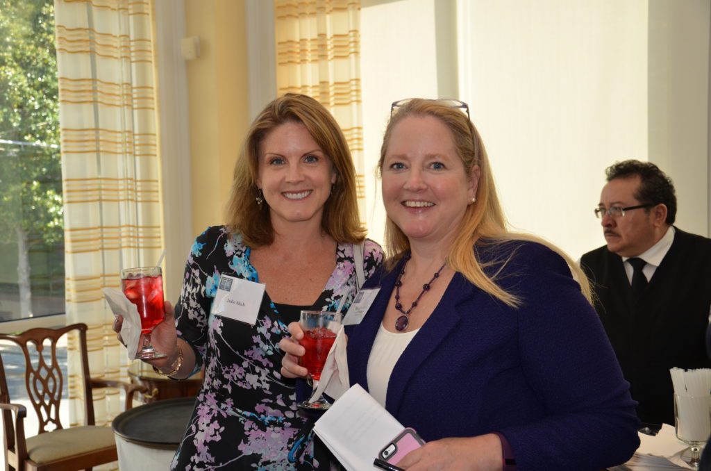 Two women smiling at a social event indoors with name tags and a drink.