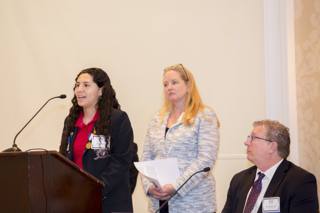 Two women and one man in a formal setting, one woman speaking at a podium.