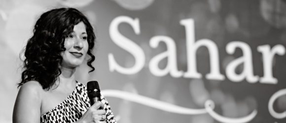 Woman speaking into a microphone at an event with 'Sah' in the background.