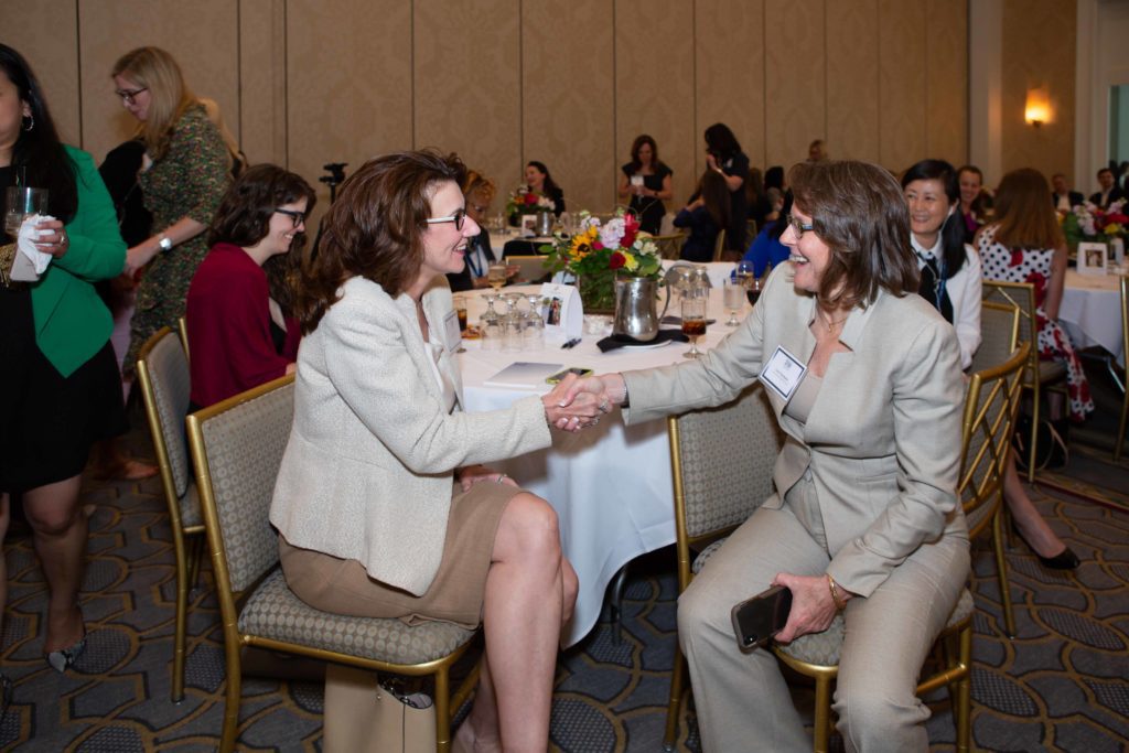 Two women warmly shaking hands while seated at a formal event table.