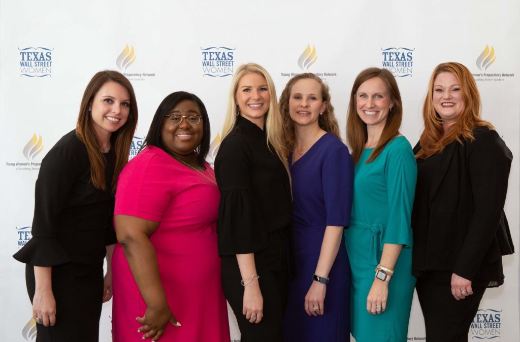 Five women standing together, smiling at the camera in a professional setting.