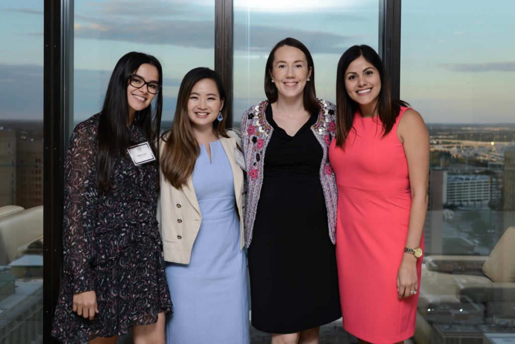 Four women smiling together indoors by large windows, dressed professionally.