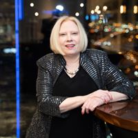 Smiling woman in a stylish jacket seated at a bar with city lights behind her.