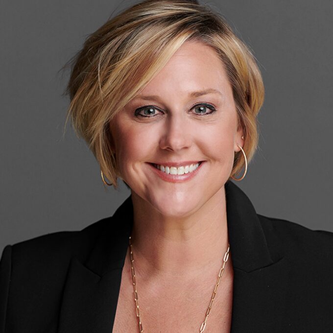 Smiling woman with short blonde hair wearing a black blazer and gold necklace.