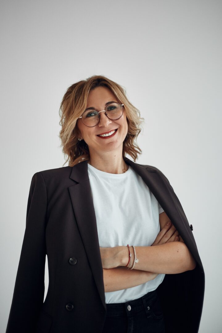 Smiling woman with glasses wearing a blazer and white shirt, arms crossed confidently.